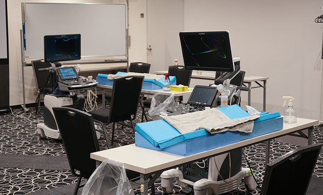 実習時の設備写真。長机の上には青い三角クッションがあり、その上にタオルが敷いてある。また、長机の先には装置が置かれている。