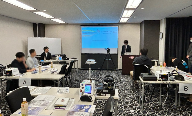 T字に組み合わされた机が3セット並んでおり、参加者が着席し講師が前で立っている会場の様子