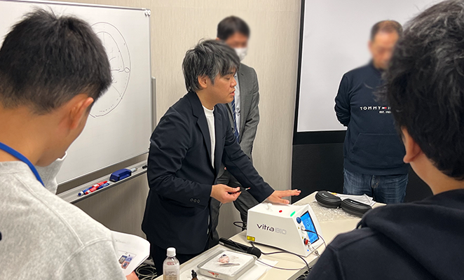 参加者に囲まれて長机の上にある機器の説明をしている仁藤稔久先生