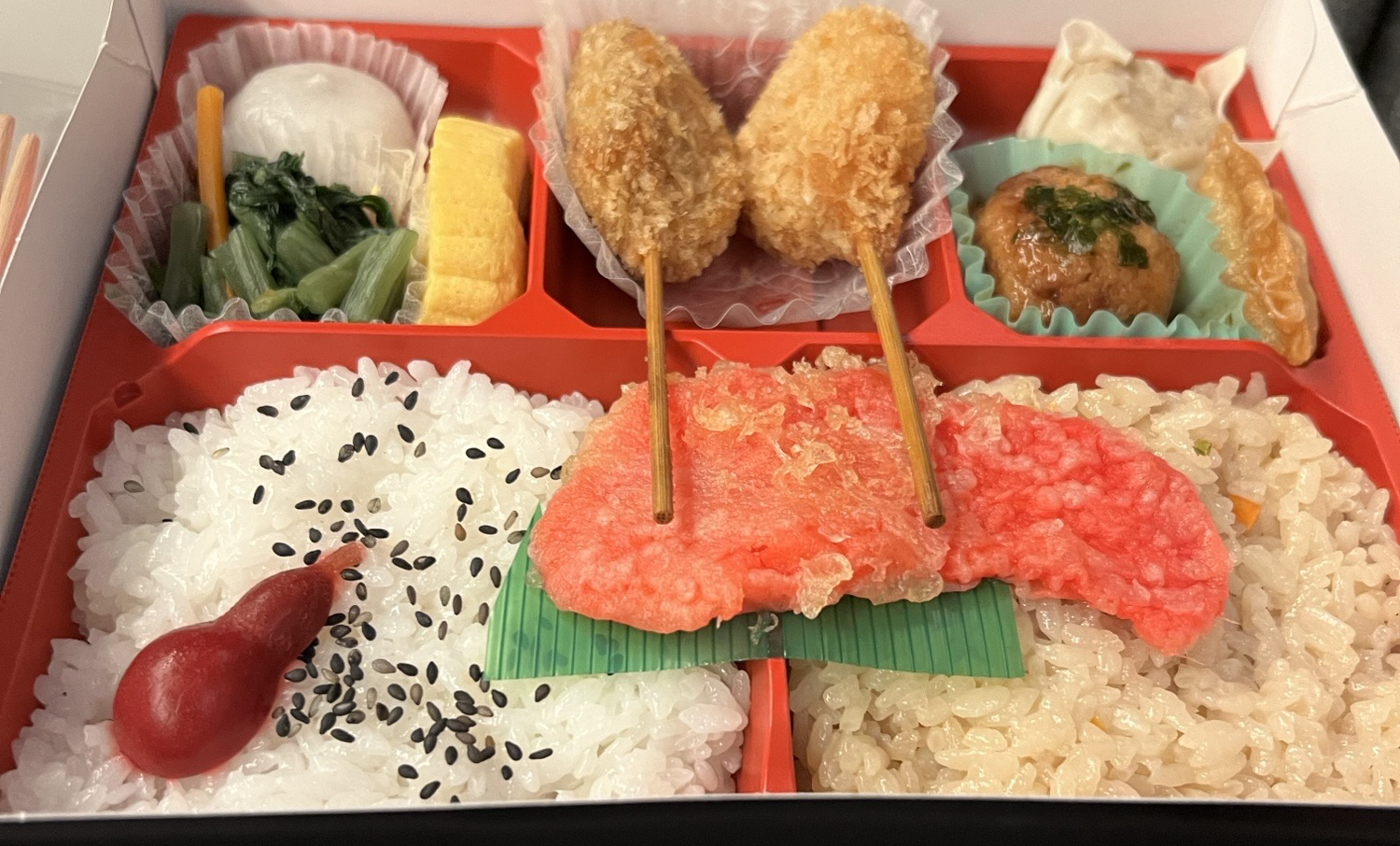 漬物、卵焼き、串カツ、シュウマイ、おこわの上に天ぷらが乗っているお弁当