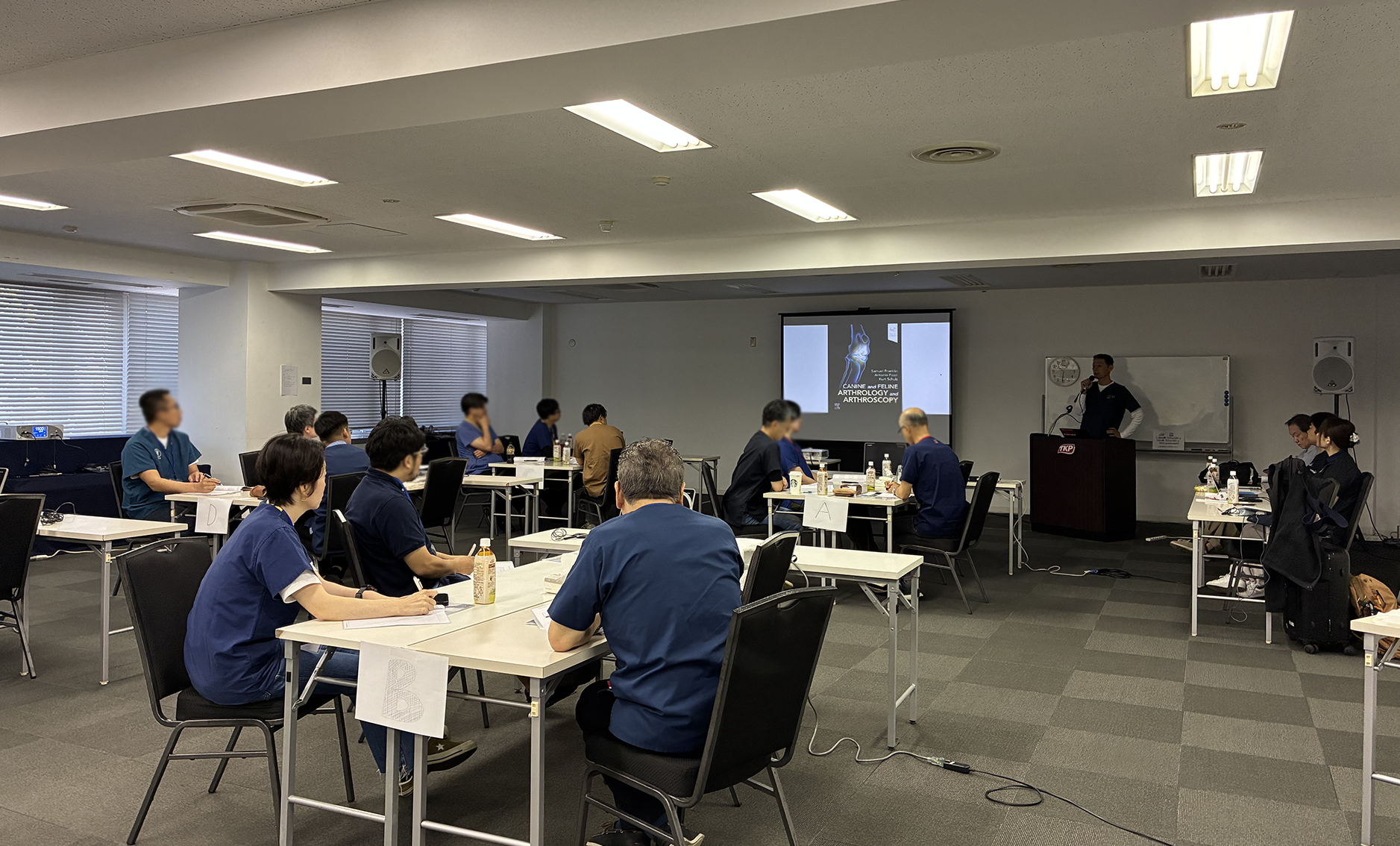 班が分かれているテーブルごとに参加者が３名ずつ着席し、正面のプロジェクタースクリーンを見ている。スクリーンの隣では講師の林先生が立って説明を行っている。